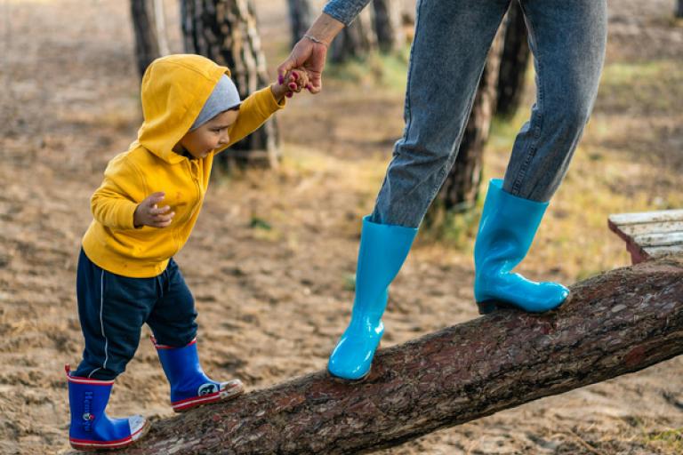 Parent holds child's hand as they walk on a log.