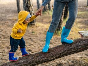 Parent holds child's hand as they walk on a log.